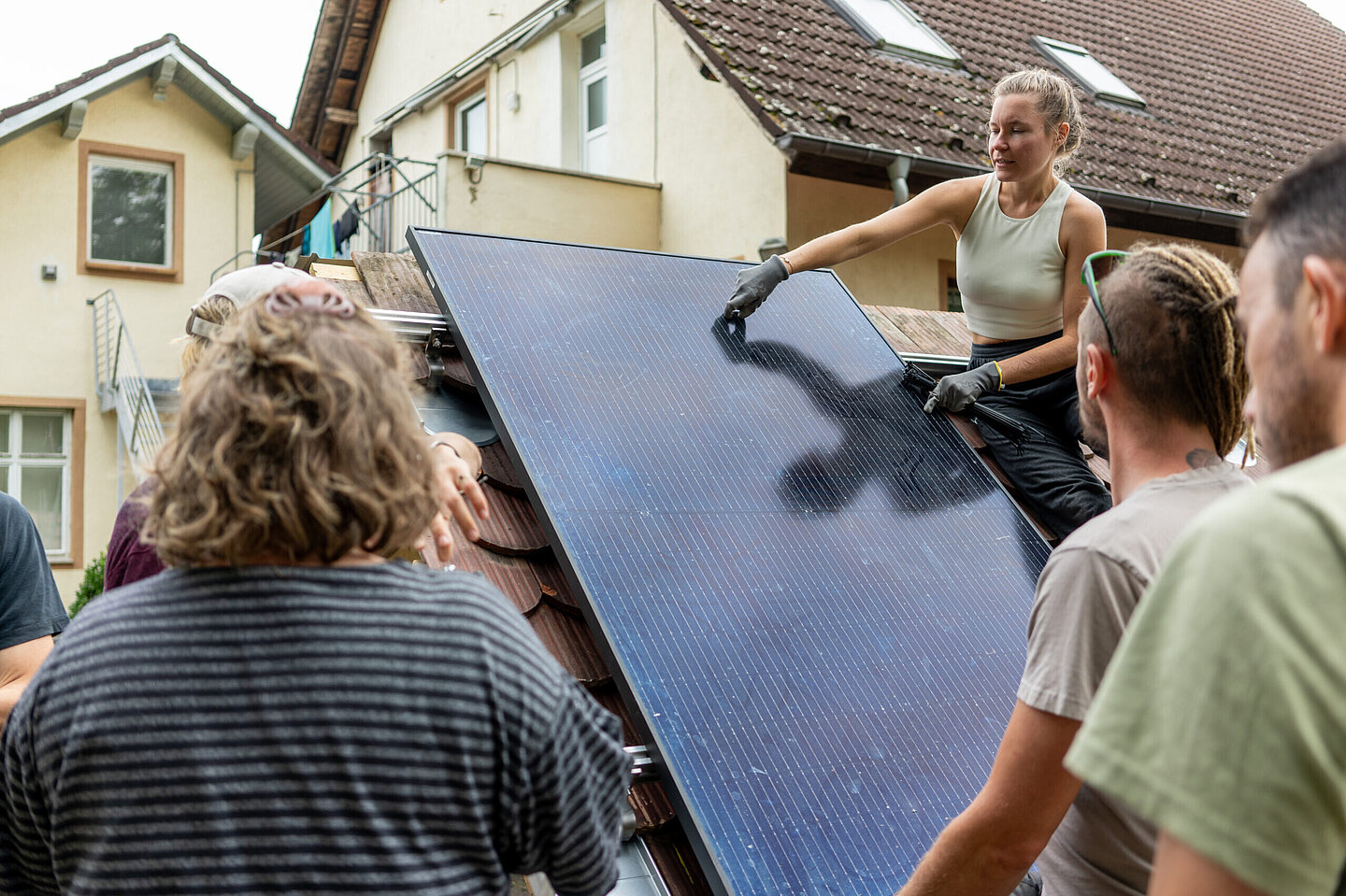 Eine Frau befestigt ein PV-Modul auf einem Dach, Personen schauen ihr zu.